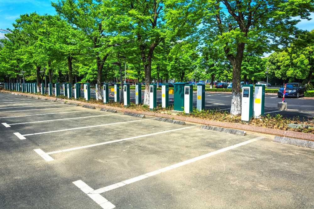 Station de recharge pour voitures électriques dans un parking ombragé par des arbres, avec de nombreuses bornes de recharge alignées et des places de stationnement vides au premier plan.